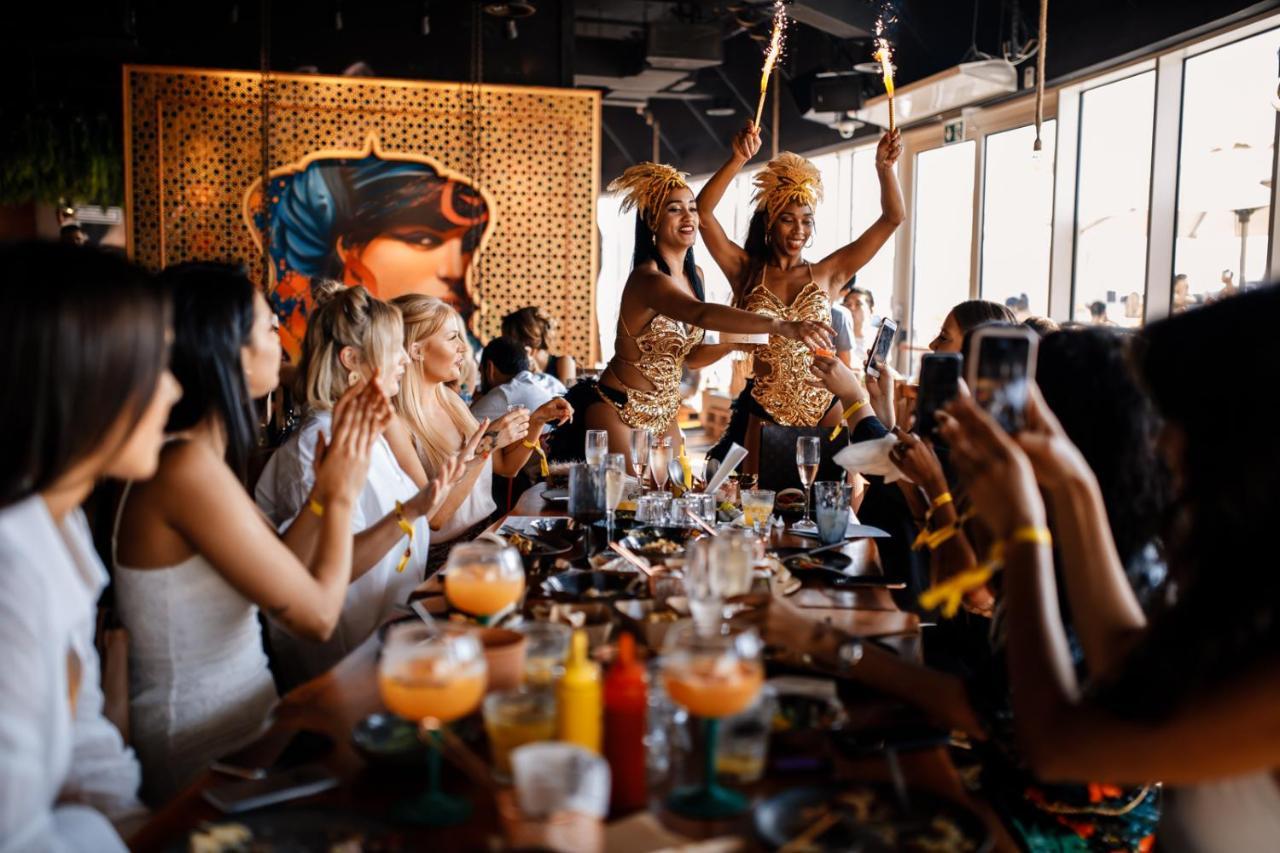 Five Jumeirah Village دبي المظهر الخارجي الصورة The photo depicts a lively restaurant scene where two performers are entertaining a group of seated diners. The performers, dressed in colorful and elaborate outfits, are holding sparklers, adding a festive atmosphere to the occasion. The diners, mos
