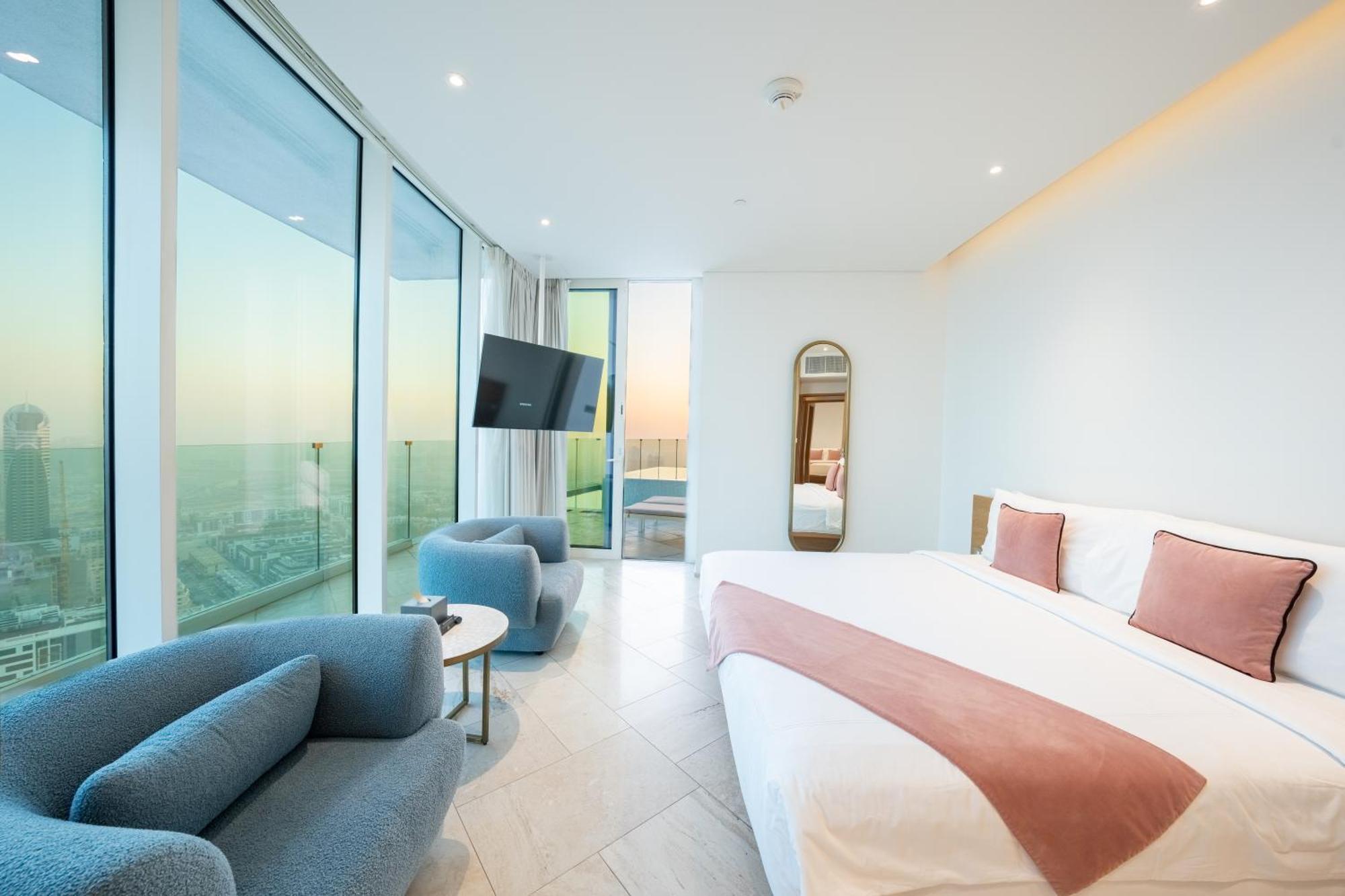 Five Jumeirah Village دبي المظهر الخارجي الصورة The photo depicts a modern hotel room featuring large windows that provide a view of the outside. The room includes a spacious bed with a pink accent blanket, a cozy blue sofa, a small round table, and a wall-mounted television. There is also a mirro